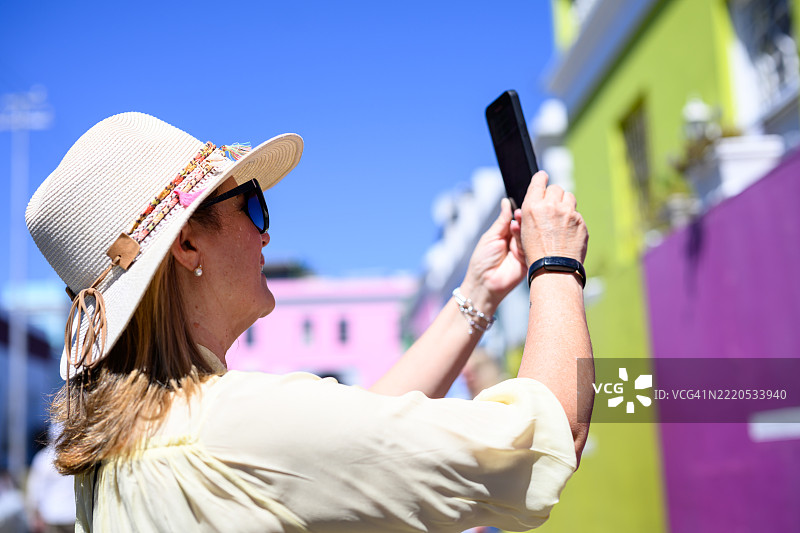 侧面特写，一位戴着时尚帽子和太阳镜的女性用智能手机拍摄色彩斑斓的房屋。图片素材