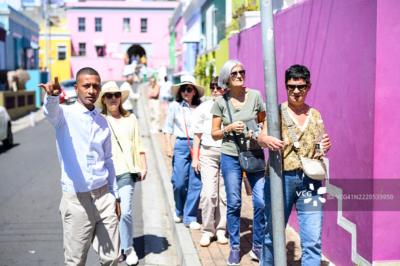 年轻的导游在阳光明媚的博卡普向一群游客指着色彩鲜艳的建筑和房屋。图片素材