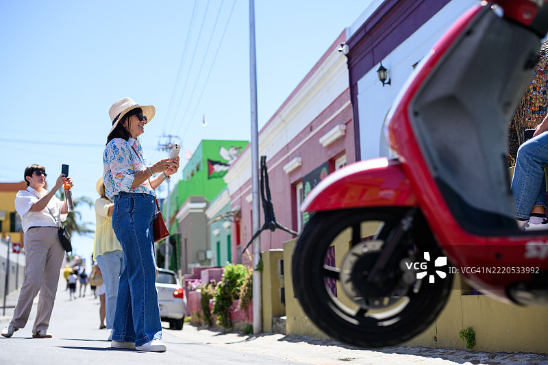 低角度拍摄的女性在色彩斑斓的街道上手持饮品，背景中有一辆红色滑板车经过。图片素材