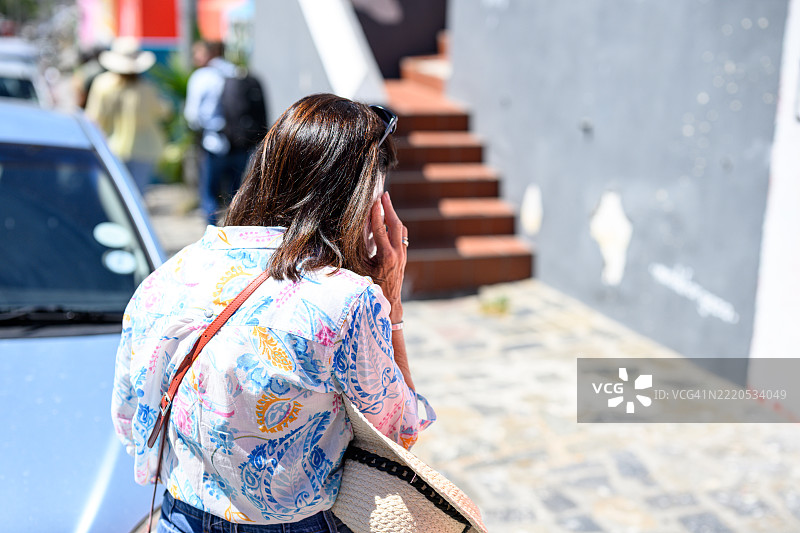 一位穿着花卉衬衫的女性在街道上站在一辆车旁接电话，背景是一栋灰色建筑。图片素材