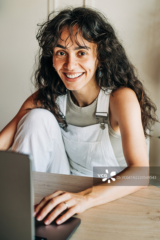一位快乐的年轻女子，留着长长的卷发，在充满植物的明亮家庭办公室里，看着笔记本电脑屏幕，脸上露出真诚的笑容。这张图片完美捕捉了远程工作、视频通话或浏览数字内容的乐趣。图片素材