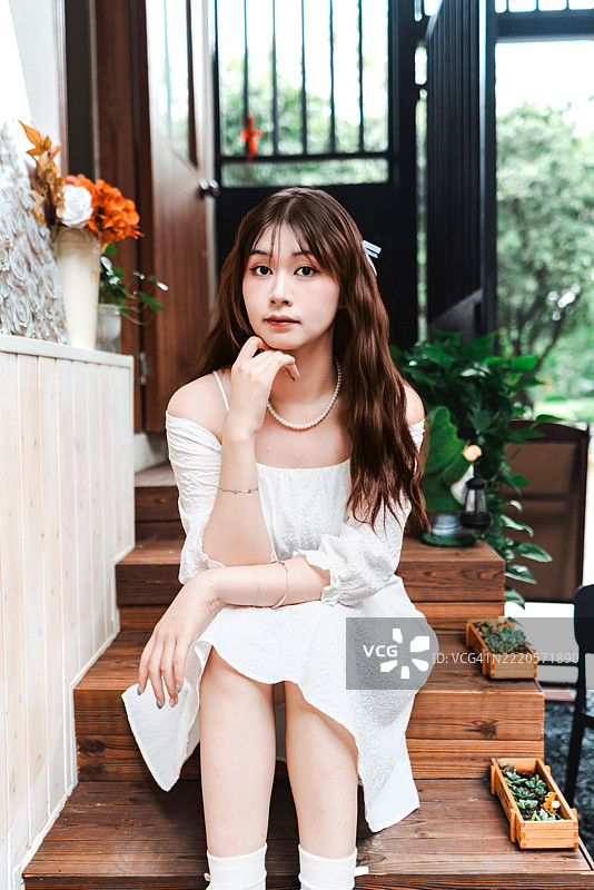 一位穿着白色连衣裙的年轻女性在花卉和木材装饰的咖啡馆入口前摆出冷静而深思的表情。图片素材