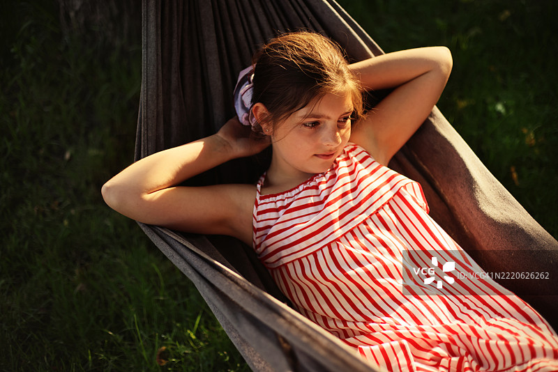 小女孩穿着红白相间的裙子，躺在花园的秋千上放松，享受日落时光。图片素材