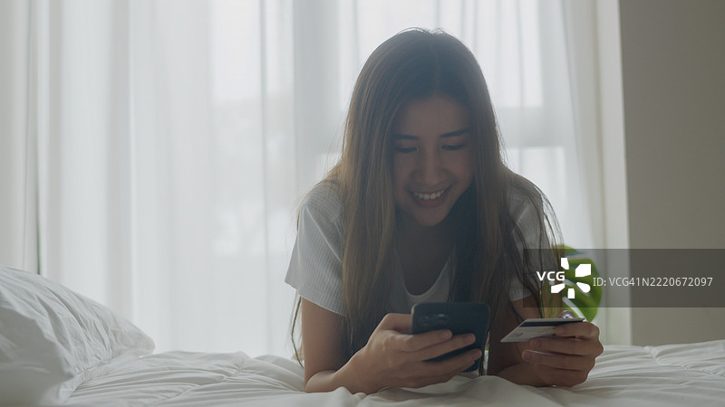 微笑的年轻亚洲女性在家床上躺着，使用信用卡和智能手机进行在线购物。电子商务、便利性和现代数字生活方式的概念。图片素材