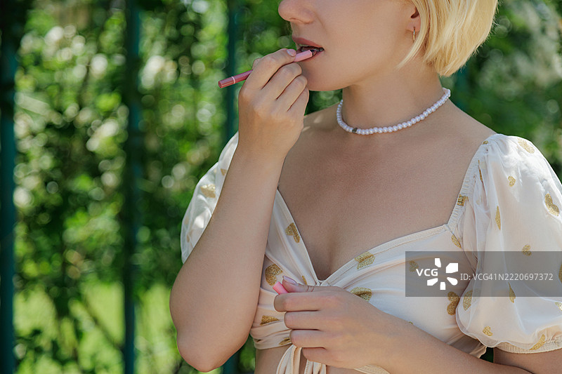 年轻的金发女性穿着白色透视上衣，在模糊的绿色植物前涂抹铅笔口红。图片素材