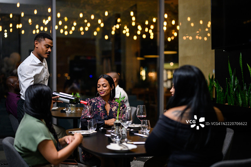男服务员正在倒红酒，一群女性在优雅的餐厅中享受晚餐，餐厅内有串灯和奢华的装饰。图片素材