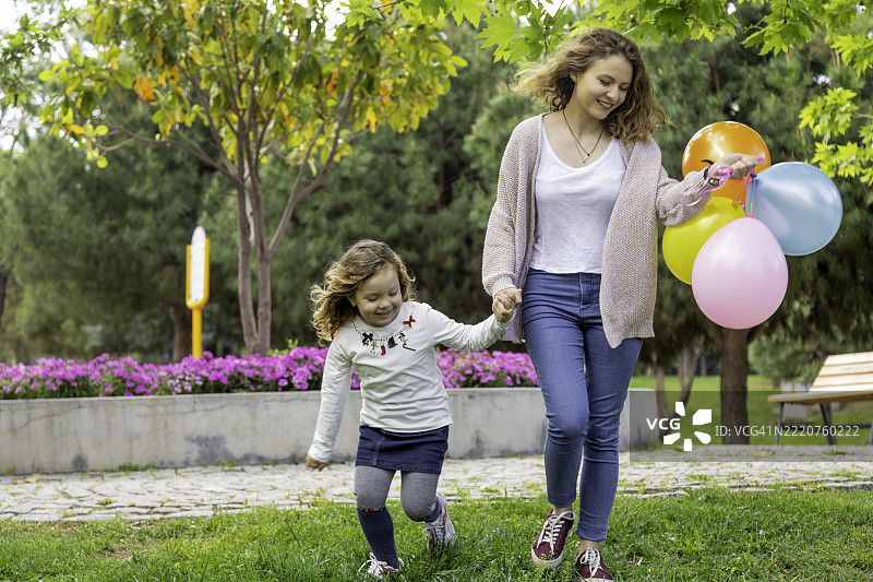 快乐的母女手牵手在绿色公园里走，手里拿着五彩缤纷的气球图片素材