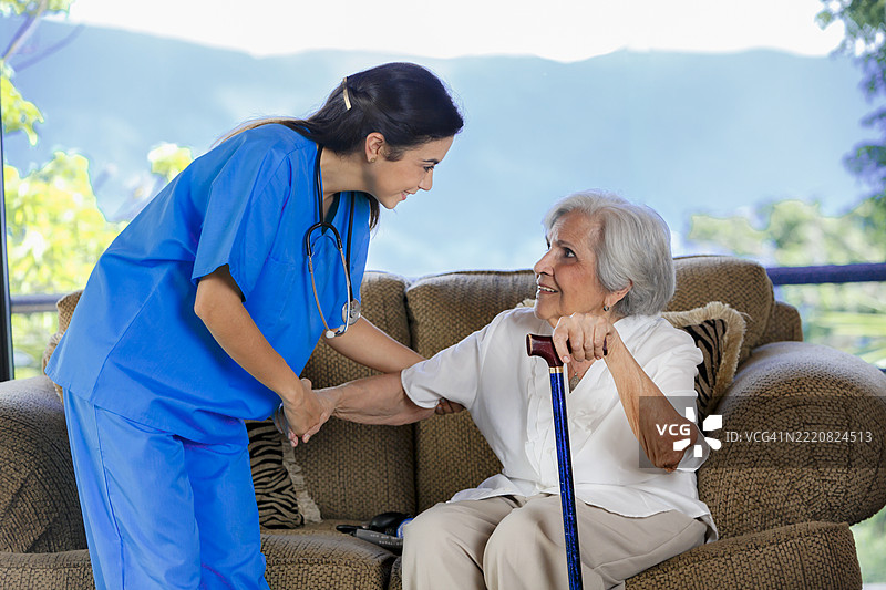 年轻的西班牙裔女性医生在家访时帮助一位老年女性站起来，提供护理院的协助。图片素材