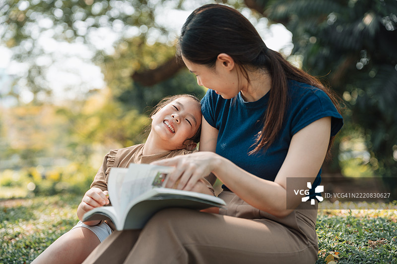 亚洲妈妈在公园的草地上给她的小女儿读书，两人坐在一起。图片素材