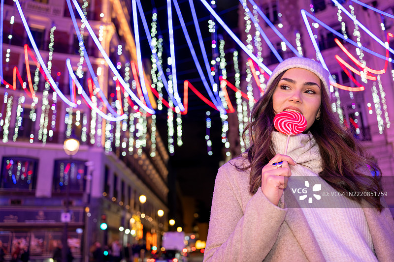 美丽的节日女性手持棒棒糖，惊叹地仰望着卡纳莱哈斯广场的圣诞灯饰和装饰。灯光璀璨的街道和欢乐的氛围展现了节日的精神。西班牙，马德里图片素材