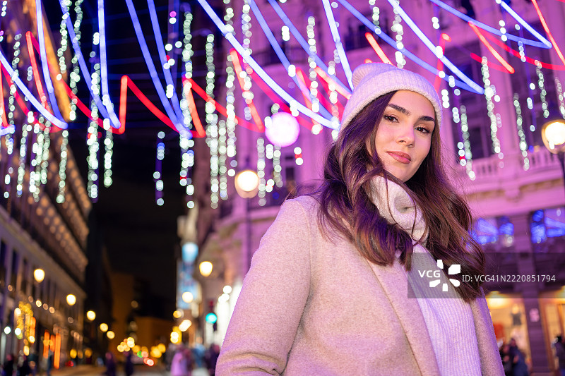 自信的白人女商人在冬季服装下，面对镜头摆姿势，身处于充满红色、蓝色和白色的璀璨圣诞灯光下，位于马德里卡纳莱哈斯广场，夜晚的圣诞假期氛围浓厚。图片素材