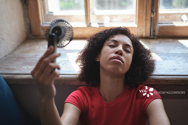 疲惫的非裔美国女性在夏季炎热中使用便携式手持小风扇，坐在窗边享受新鲜空气，在家中降温度过热浪。图片素材