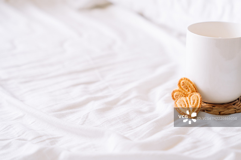 白色咖啡杯特写，旁边有两个心形饼干，放在编织的杯垫上， resting on wrinkled white sheets. 简约的床上早餐，温馨宁静的家居时刻。图片素材