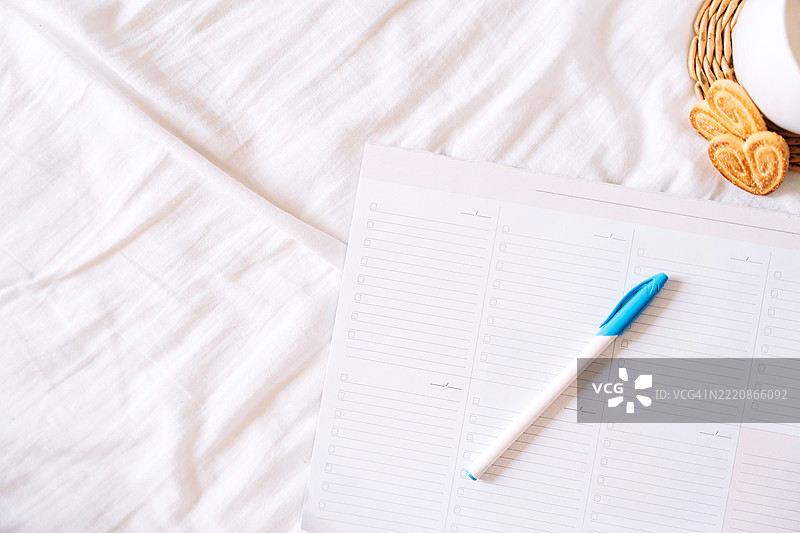 日常计划本、钢笔、卡布奇诺和饼干平铺在藤编杯垫上，背景为白色布料。明亮的极简美学，适合规划、设定目标和享受宁静的早晨。图片素材