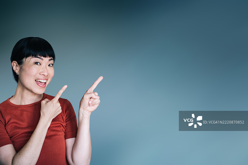 穿着红色衬衫的快乐亚洲女性，双手食指指向上方，面带灿烂笑容，背景为蓝色。图片素材