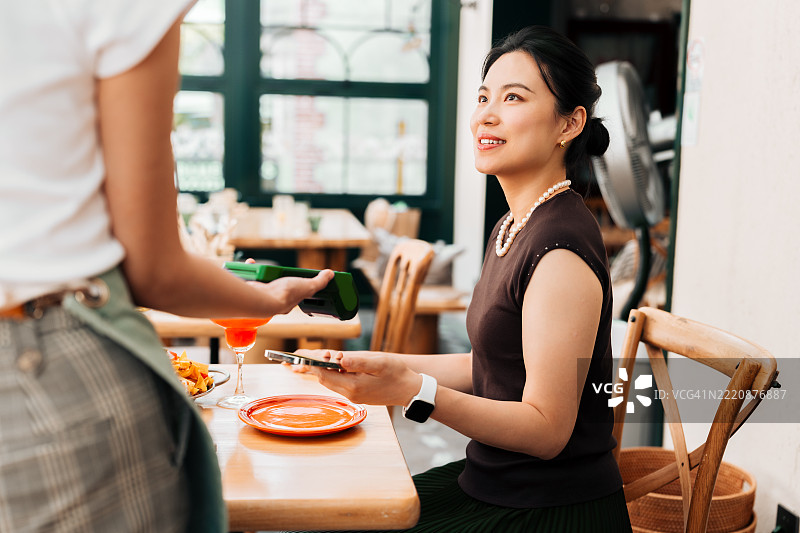 年轻美丽的亚洲女性在餐厅侧面视图中使用智能手机进行移动支付，扫描并在刷卡机上支付账单，快速便捷地完成非接触式支付。NFC技术，轻触即付的概念。图片素材