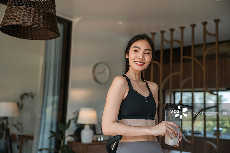 运动型年轻女性在家中骑健身车喝乳清蛋白饮料的肖像图片素材