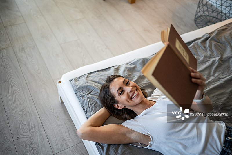 年轻女性躺在床上阅读一本书，面带微笑图片素材