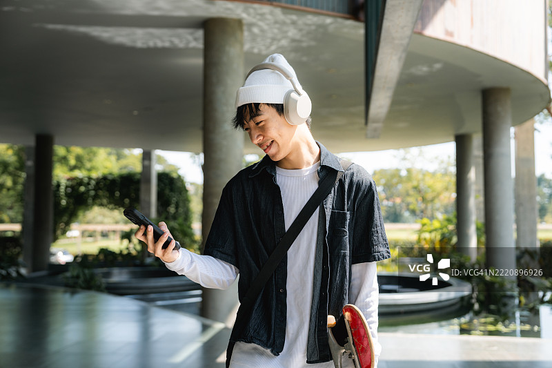 微笑的大学生在校园里走着，手持数字平板，戴着耳机听音乐。图片素材