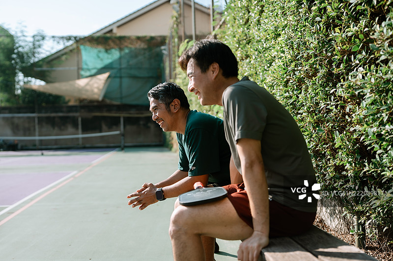 两个快乐的成熟男性在球场边坐着，经过一场热烈的匹克球比赛后，进行着真诚的交谈，互相了解对方的生活。图片素材