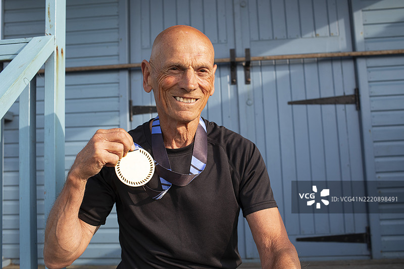 一位老年男子坐在海滩小屋旁，手里拿着一枚跑步奖牌图片素材