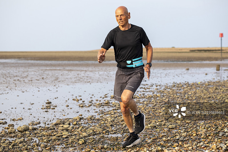 活跃的老年男子在退潮时沿着卵石海滩的海岸线跑步图片素材