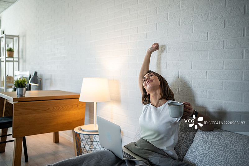年轻女性在床上醒来并伸展身体，旁边有咖啡和笔记本电脑图片素材