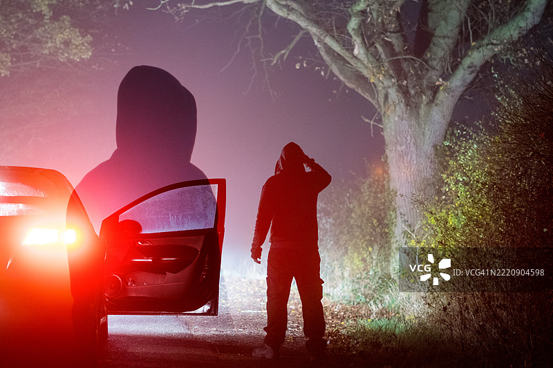 一个恐怖概念的阴森鬼魂双重曝光的披风人物，站在一辆停着的车旁。在乡村的阴森孤独的雾霭夜路上。图片素材