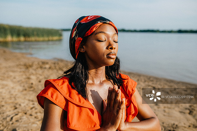 一位女性站在海滩上,双手合十,穿着鲜艳的红色服装和图案头巾。图片素材