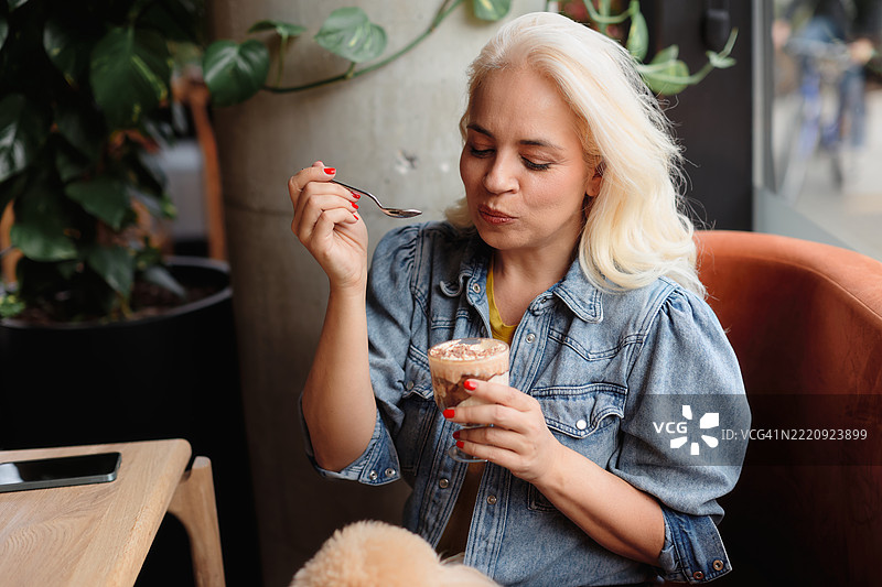 一位五十多岁的美丽女性坐在咖啡馆的窗边，享受着提拉米苏蛋糕。图片素材