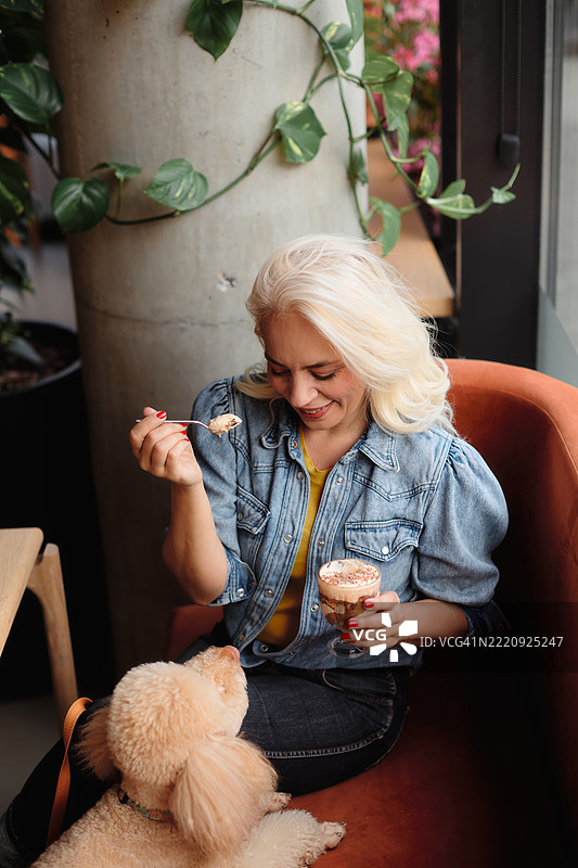 一位五十多岁的美丽女性坐在咖啡馆的窗边，享用她的提拉米苏蛋糕，旁边坐着她的棕色贵宾犬。图片素材