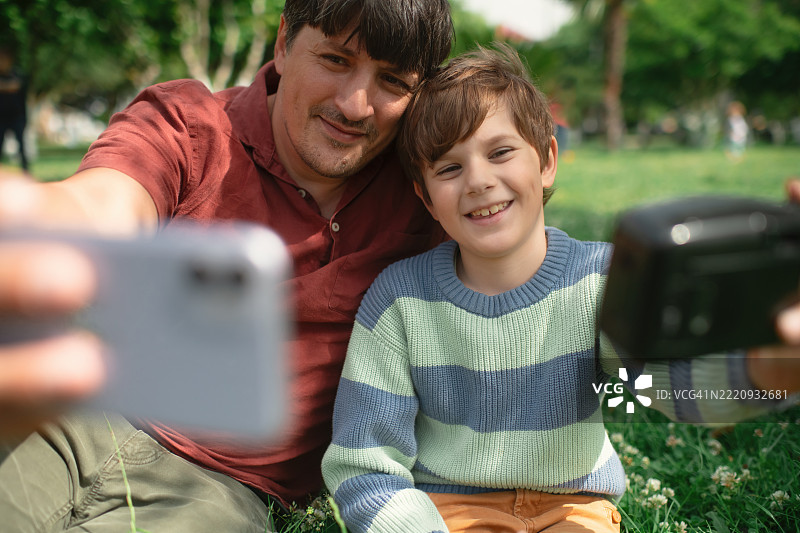 小男孩和他的父亲在公园里用智能手机和复古胶卷相机拍照图片素材