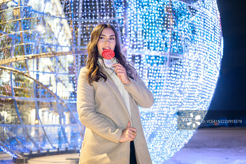 快乐的年轻女性在西班牙广场（Plaza de España）的大圣诞球前享受五彩缤纷的棒棒糖，周围是节日灯光照亮的寒冷冬夜。马德里，西班牙图片素材