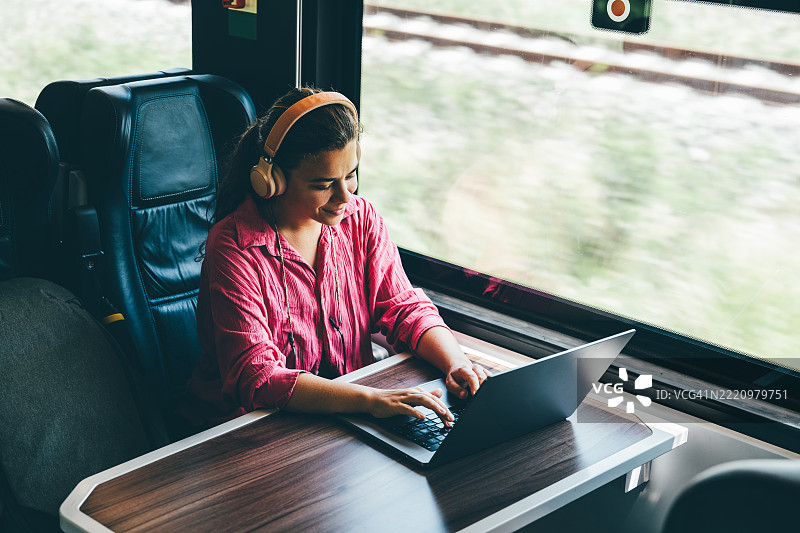 一位戴着耳机、身穿便服的年轻女子正坐在火车上，靠着窗户，专心致志地使用笔记本电脑工作。她专注而放松的表情，恰如其分地展现了当今人们在旅途中进行远程办公、学习和高效工作的现实状态。图片素材