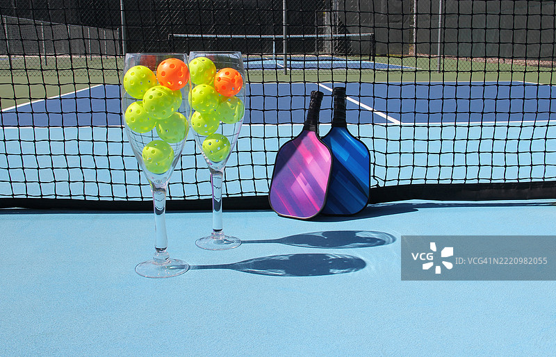 巨大的酒杯里装满了乒乓球，放在户外的乒乓球场上，网附近有两只球拍。图片素材