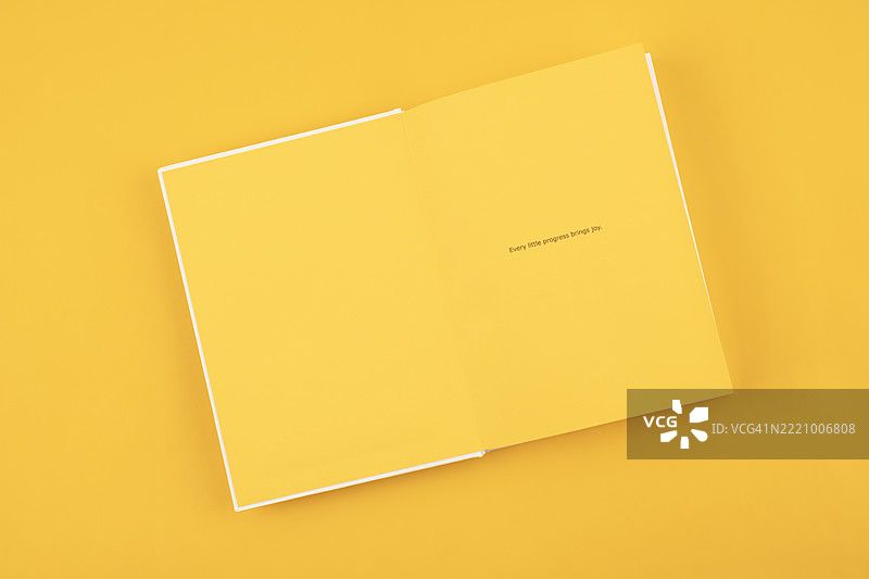 从上方俯视打开的黄色记事本，背景为黄色，上面写着“每一点进步都带来快乐”的文字。图片素材
