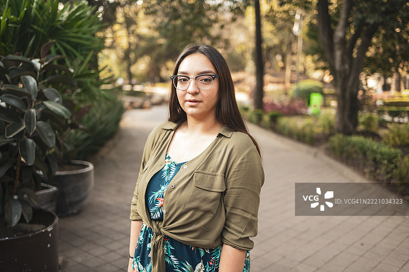 年轻女性穿着休闲服装，站在阳光明媚的公园里，正看着镜头。图片素材