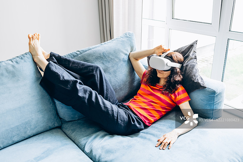 年轻女性在家沙发上使用虚拟现实头盔。日常生活中的虚拟现实，家庭娱乐与VR。图片素材
