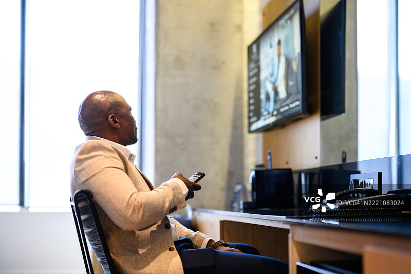 光头商人在现代酒店房间内手持遥控器观看电视，窗外光线明亮。图片素材