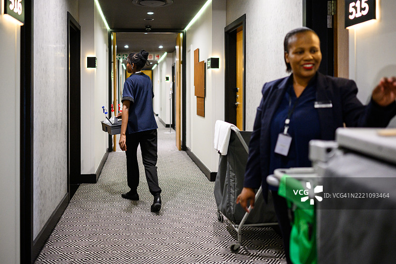 快乐的酒店主管在清洁推车旁工作，女佣沿着走廊走开。图片素材