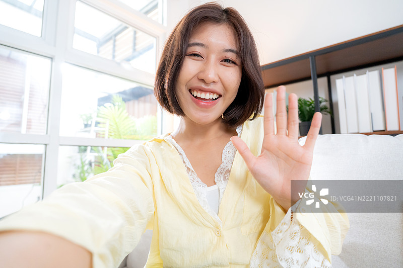 年轻的亚洲女性在家中的客厅里挥手微笑，享受在线视频通话，友谊与沟通，数字生活方式，科技与社交连接，科技视角。图片素材