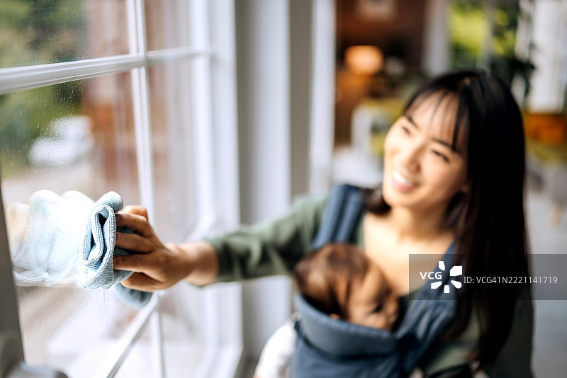 女人一边清洁窗户，一边抱着她可爱的小女儿在婴儿背带里。图片素材