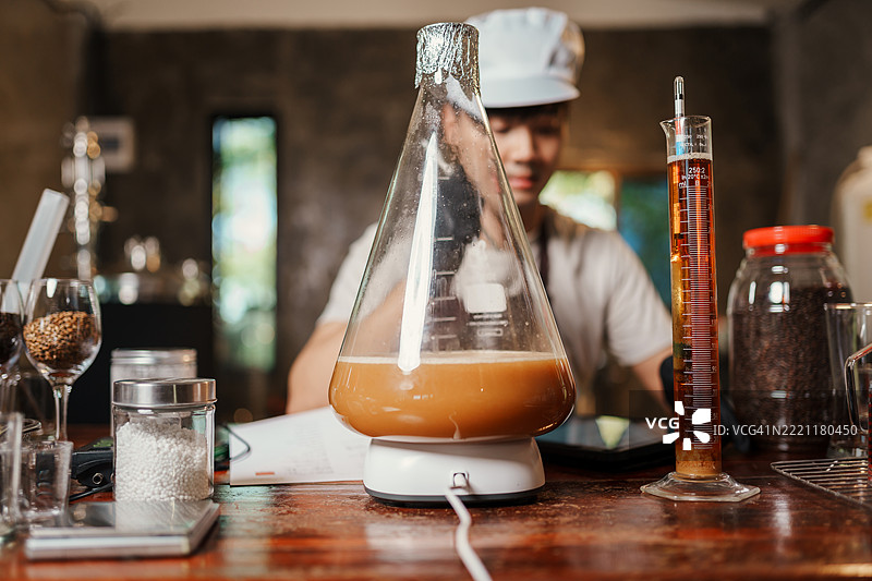 活跃的酵母发酵液在磁力搅拌器上的烧瓶中旋转的特写，亚洲家庭酿酒师在记录酿造数据。图片素材