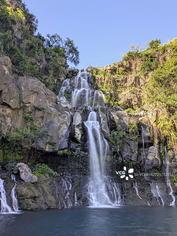 一座高耸的瀑布从岩石悬崖倾泻而下，周围绿树成荫，蓝天清澈，位于法国留尼旺岛，非洲。图片素材