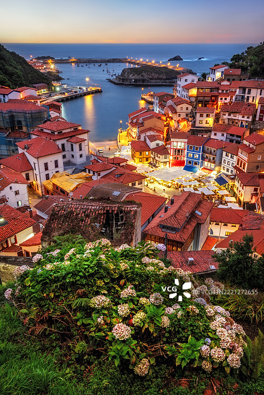 五彩斑斓的房屋沿着阿斯图里亚斯西班牙库迪列罗的阶梯状街道倾斜而下，在蓝色时刻熠熠生辉，前景中盛开的花朵增添了生机。图片素材