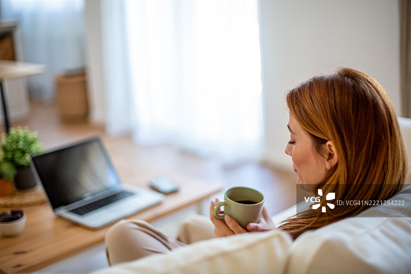 女人在客厅的沙发上放松，手里拿着咖啡和笔记本电脑。图片素材