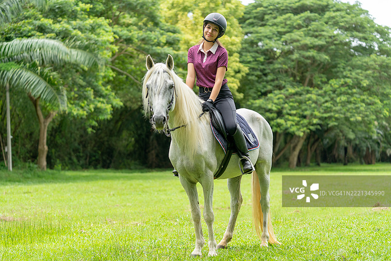 可爱的少女坐在马背上，微笑着并看向镜头，在农场或公共花园中放松，背景是一棵大树。图片素材