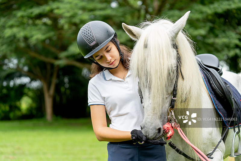年轻的少女穿着白色衣服，站在白马面前，给它喂食，她们在农田或公共花园的草地上，神情放松。图片素材
