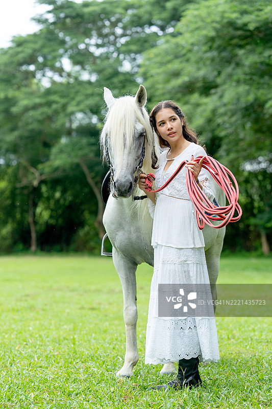 年轻女孩穿着白色连衣裙，站在白马面前，指向左侧，背景为农场或公共花园。图片素材