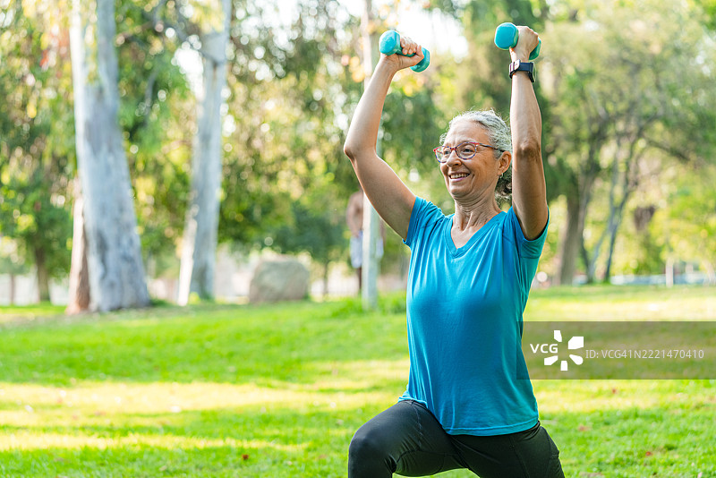 微笑的老年女性在公园举重，锻炼肌肉和力量，使用哑铃或进行无氧运动。退休后的活力与健康。健美的女性正在训练。图片素材
